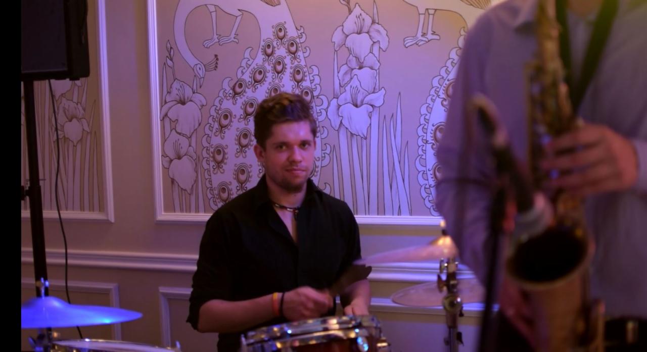 Perkusista z pałeczkami i saksofonista na tle ściany z motywem pawich piór podczas występu.