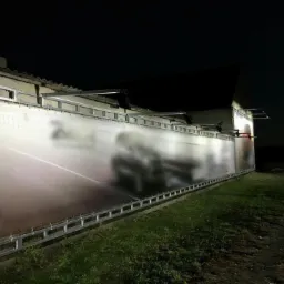 Oświetlony baner reklamowy zamontowany na elewacji budynku, widoczny w nocy przy słabym oświetleniu, z zaparkowanym samochodem obok.