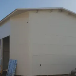 Nowo wybudowana, jasna hala z płyty warstwowej, z dwoma otwartymi wjazdami, widoczne elementy konstrukcyjne dachu, częściowo zasłonięta przez żółty podnośnik i zaparkowany samochód, w tle błękitne...