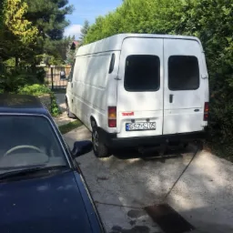 Biały dostawczy samochód marki Transit zaparkowany na podjeździe, widok z tyłu, z widoczną tablicą rejestracyjną GZSZ S205, częściowo zasłonięty przez ciemny samochód osobowy.