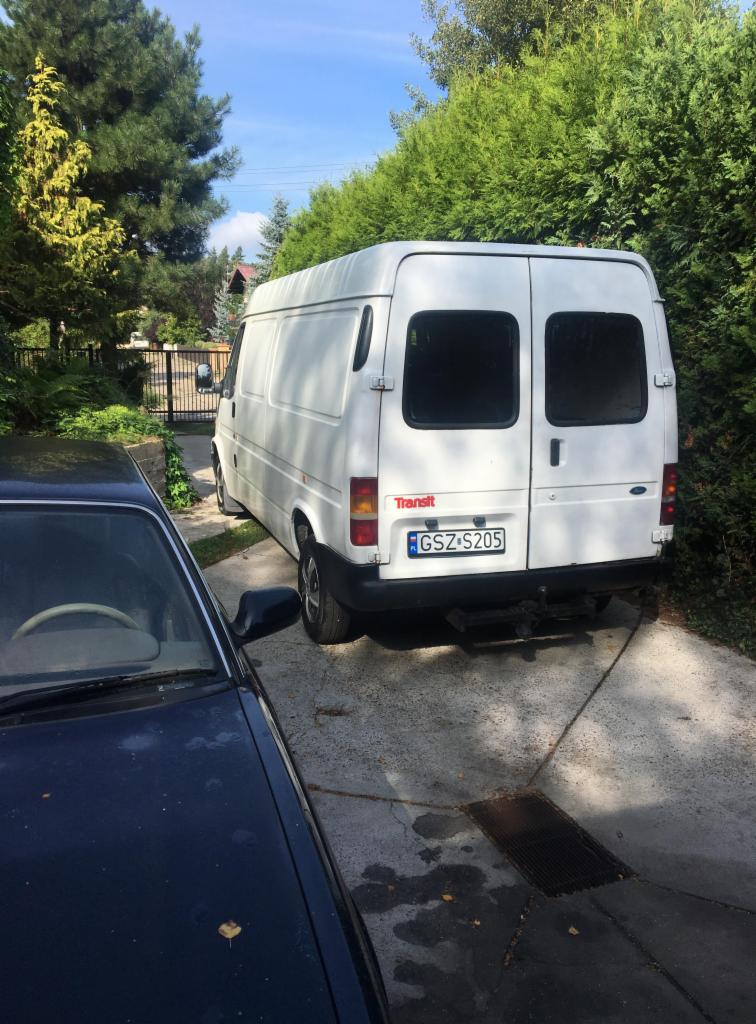 Biały dostawczy samochód marki Transit zaparkowany na podjeździe, widok z tyłu, z widoczną tablicą rejestracyjną GZSZ S205, częściowo zasłonięty przez ciemny samochód osobowy.