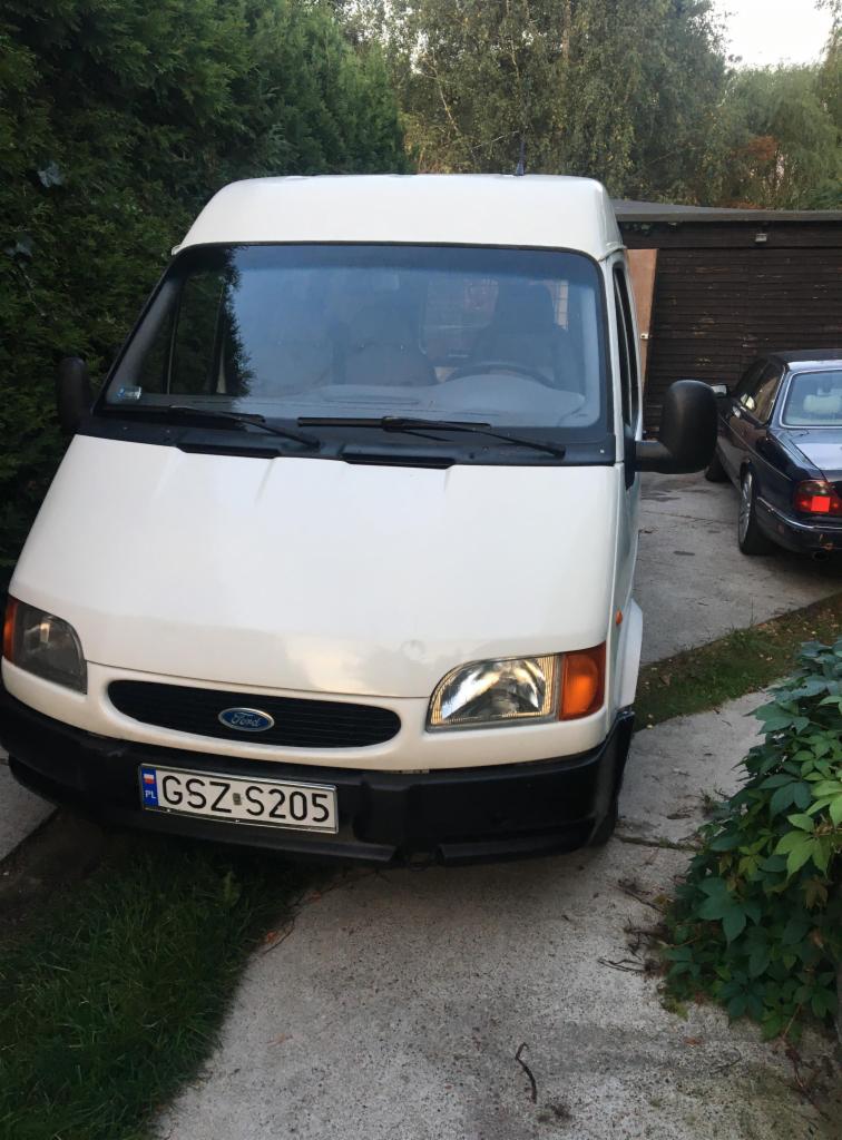 Biały Ford Transit zaparkowany na betonowym podjeździe, widok z przodu, z widoczną tablicą rejestracyjną i fragmentem ciemnego samochodu w tle.