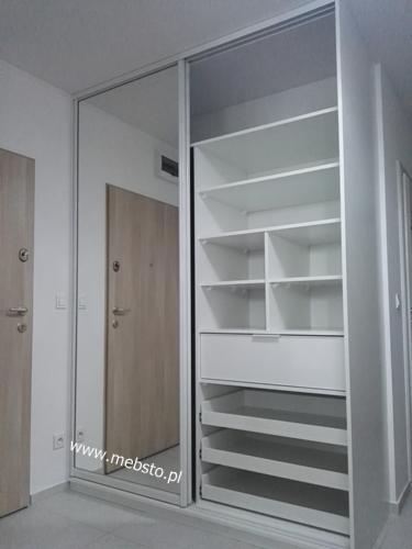 Biała szafa wnękowa z przesuwnymi drzwiami, z lustrem na jednym skrzydle, widoczne półki i szuflady, minimalistyczny design.