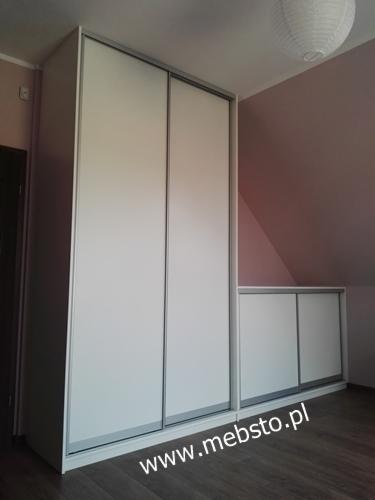 Biała szafa z przesuwnymi drzwiami, dopasowana do skosu poddasza, z widocznym oświetleniem sufitowym i adresem strony internetowej na podłodze.