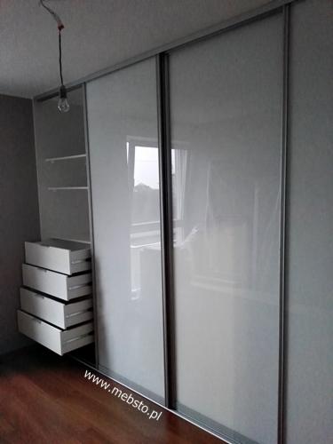 Biała szafa wnękowa z przesuwnymi drzwiami, widoczne półki i szuflady, surowy wystrój wnętrza, żarówka na kablu zwisa z sufitu.