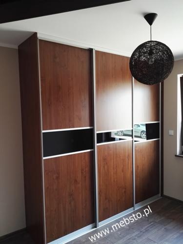 Trzydrzwiowa szafa z okleiną drewnopodobną, czarnymi wstawkami i lustrzanymi fragmentami, oświetlona dekoracyjną lampą wiszącą.