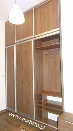 Wysoka szafa z przesuwnymi drzwiami w odcieniach drewna, z otwartą wnęką z półkami na buty i wieszakami na ubrania, oświetlona wiszącą lampą.
