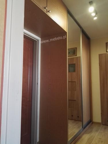 Wnętrze szafy wnękowej z przesuwnymi, lustrzanymi drzwiami, widoczna również część futryny drzwi wejściowych, naklejka z adresem strony internetowej na boku szafy.