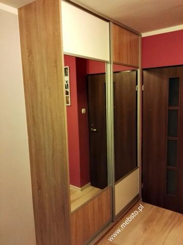 Szafa wnękowa z przesuwnymi drzwiami, kombinacja drewna i białych paneli, widoczna w odbiciu część pokoju z czerwonymi ścianami i drewnianymi drzwiami.