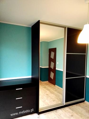 Czarna szafa z przesuwnymi drzwiami z lustrem, obok czarna komoda z trzema szufladami, widoczna w lustrze drewniana drzwi z szybami, ściany w kolorze turkusowym.