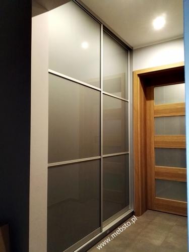 Wysoka szafa z przesuwanymi drzwiami z matowego szkła i aluminiowymi ramami, widoczna obok drewnianych drzwi z czterema szklanymi panelami w korytarzu.