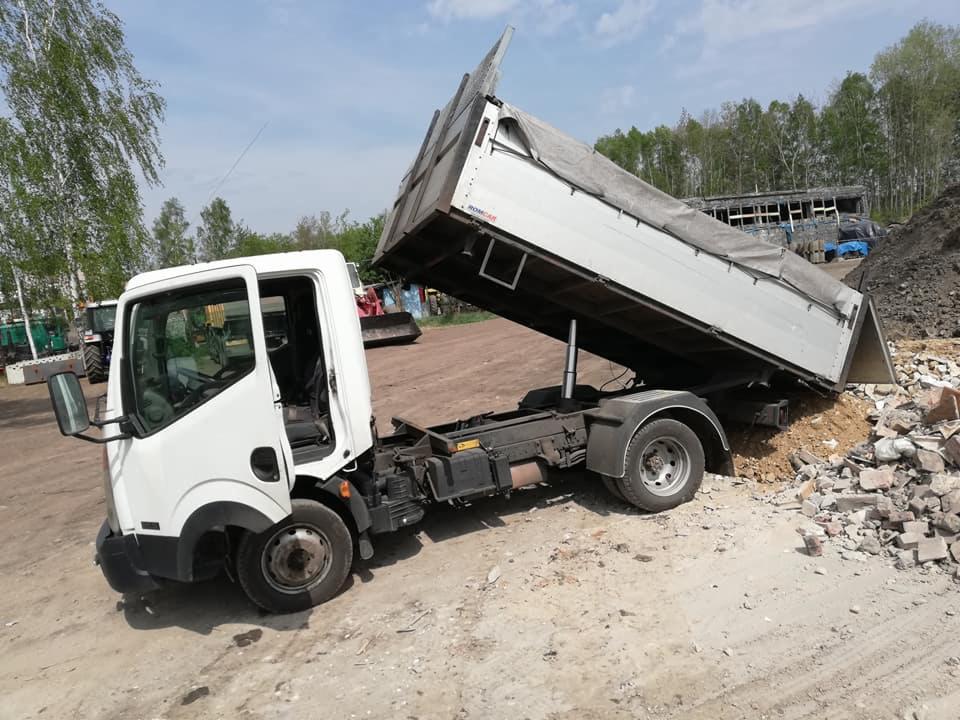 Biała ciężarówka wywrotka z podniesioną skrzynią ładunkową wysypująca gruz na placu budowy, na tle drzew i budynku w oddali.