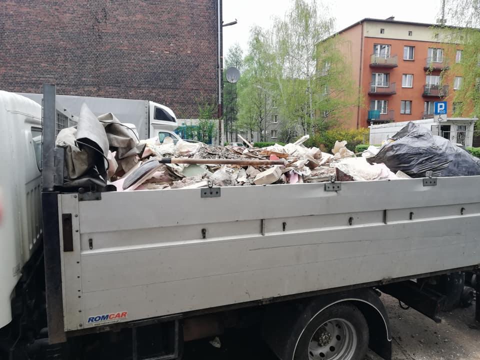 Biała ciężarówka Romcar wypełniona gruzem, fragmenty cegieł i odpadów budowlanych, widoczna łopata i czarny worek na tle budynków mieszkalnych.