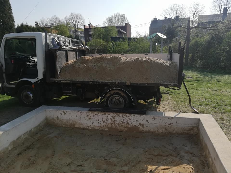Ciężarówka z otwartą kabiną, wypełniona piaskiem, obok świeżo wybudowanej piaskownicy na trawniku w słoneczny dzień.