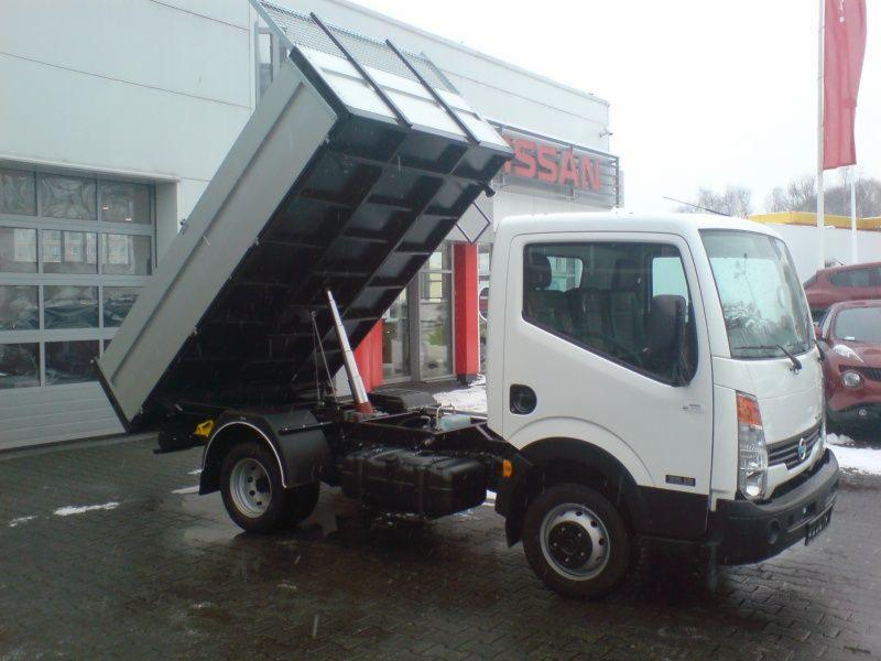 Biały samochód dostawczy Nissan z otwartą skrzynią ładunkową typu wywrotka, zaparkowany na mokrym placu przed salonem samochodowym.