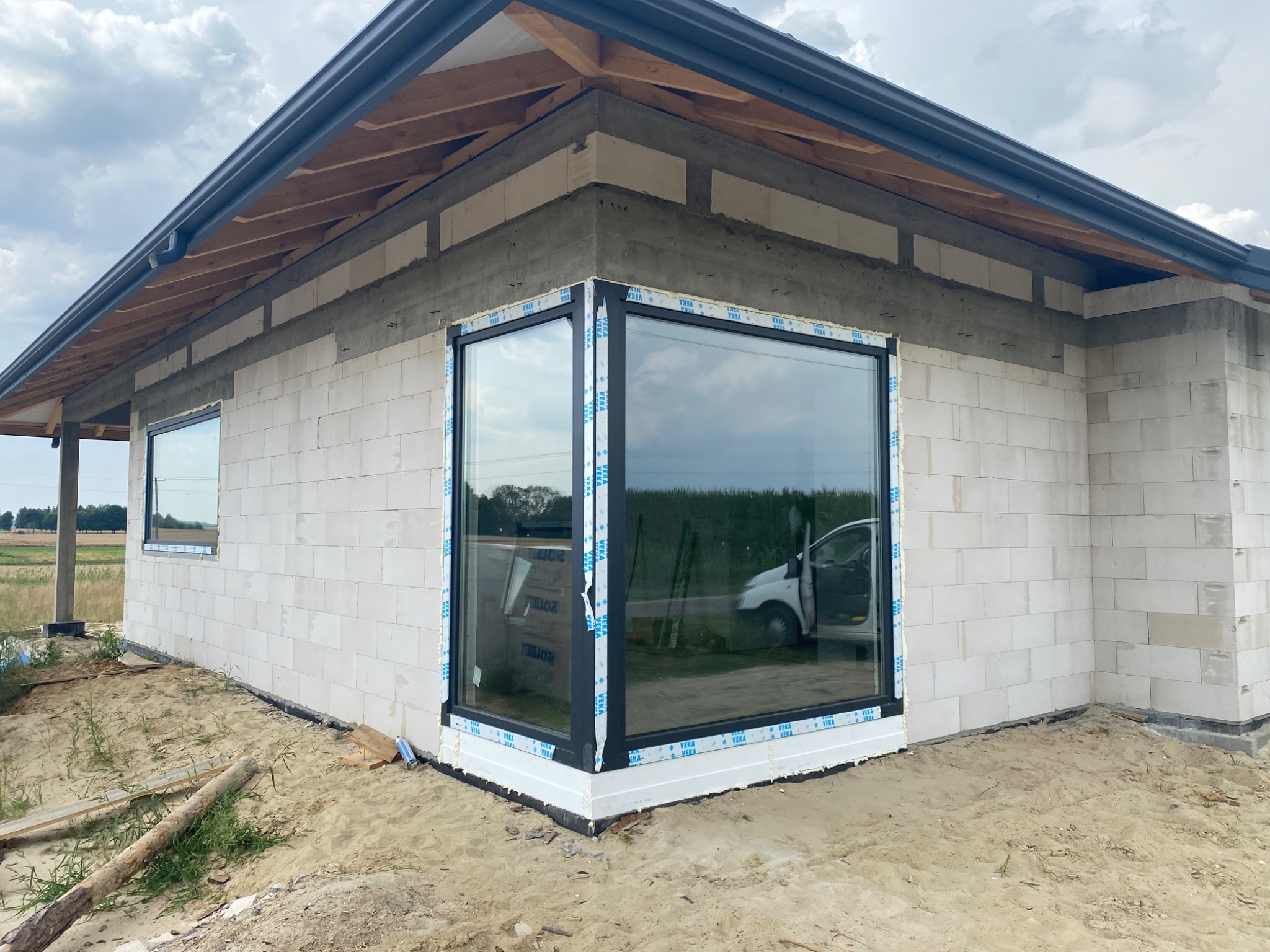 Narożne okno z czarną ramą zamontowane w surowym budynku z białych bloczków, widoczna folia ochronna na ramie i samochód w odbiciu szyby.