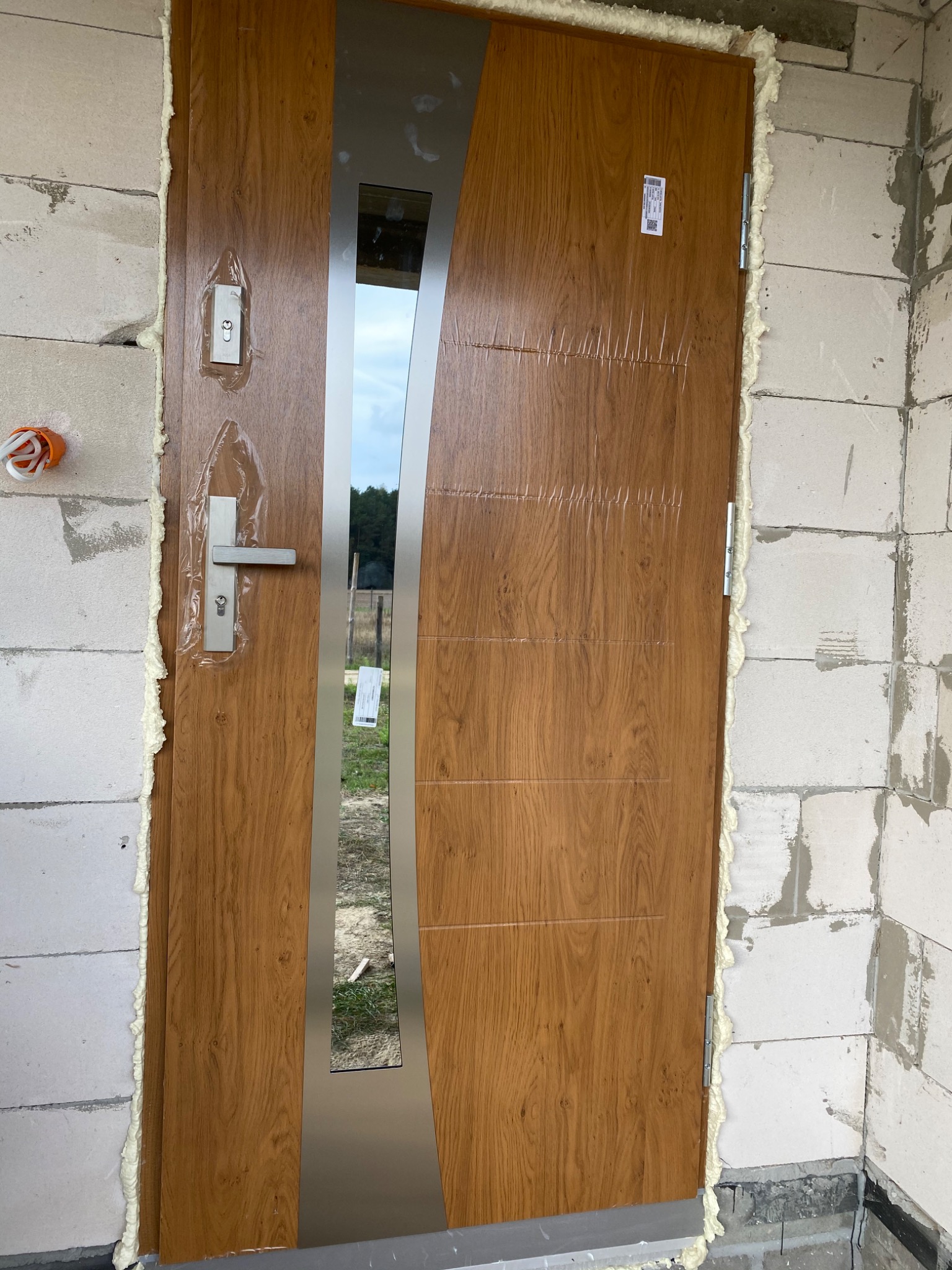 Montaż brązowych drzwi zewnętrznych z metalową wstawką i ościeżnicą, zabezpieczonych folią ochronną, pianka montażowa wokół ościeżnicy, ściany z betonu komórkowego.