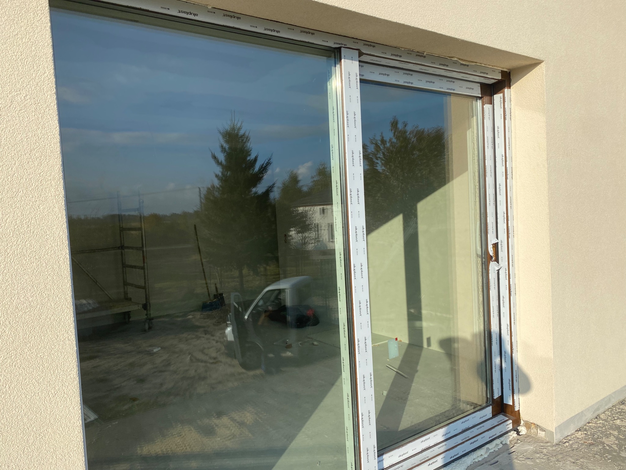 Nowo zamontowane okno przesuwne z PCV, z widocznym odbiciem krajobrazu, w tym drzewa i samochodu, na tle jasnej elewacji budynku.