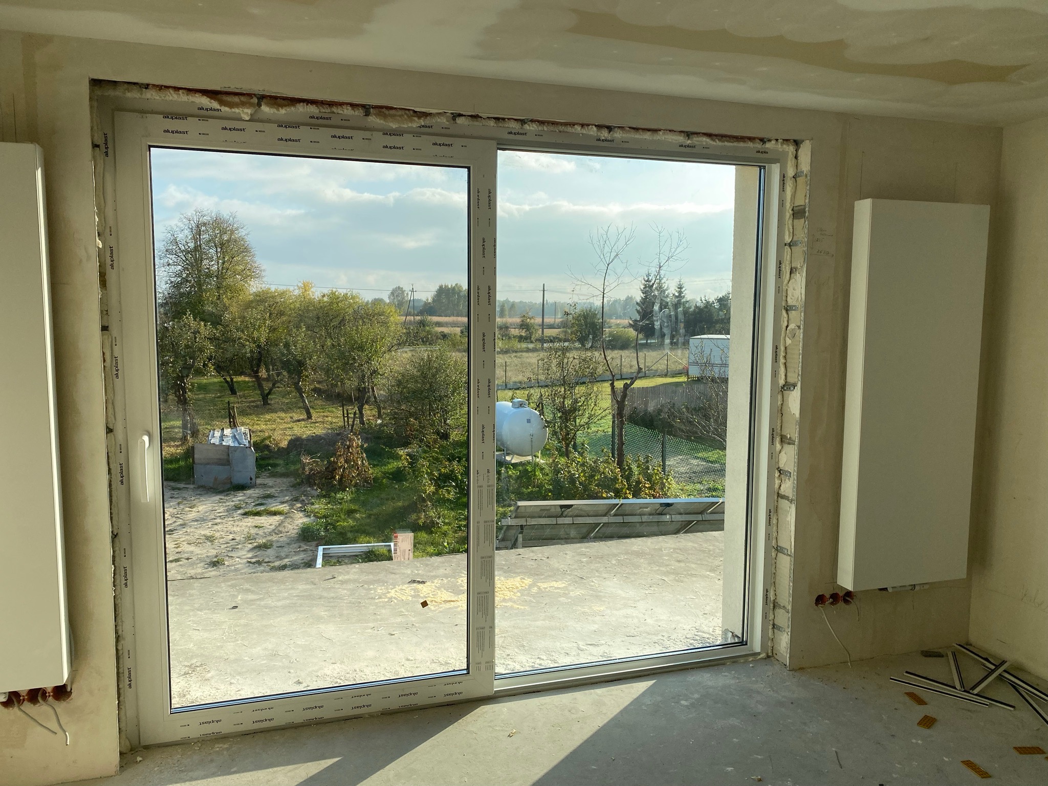 Montaż okna przesuwnego z PCV, widok na zewnątrz, surowy stan wykończenia wnętrza, pianka montażowa widoczna wokół ramy, elementy montażowe na podłodze.