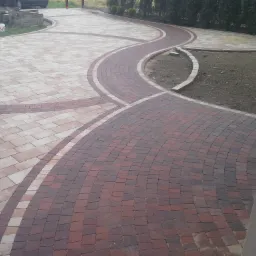 Ukończony podjazd z kostki brukowej w dwóch odcieniach, ułożonej w falujący wzór z widocznym zapasem kostki i obrzeżami trawnika.