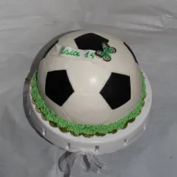 Tort w kształcie piłki nożnej z zielonym lukrem imitującym trawę, dekorowany motylkiem i napisem 'Asia 14' na białej wstążce.