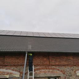 Paweł Romańczyk - Instalator na drabinie montuje panele słoneczne na dachu budynku z cegły, widoczne rynny.
