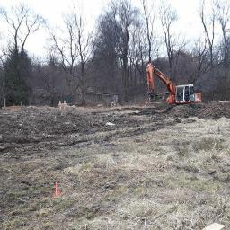 Michał Stanisławczyk BSGEO - Pomarańczowa koparka Hitachi na placu budowy z wykopanymi fundamentami i drewnianymi stemplami, widok na zalesiony teren.