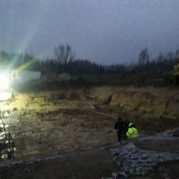 Michał Stanisławczyk BSGEO - Prace geodezyjne na placu budowy po zmroku, widoczny oświetlony wykop, dwaj geodeci w kurtkach oraz koparka CAT w tle.