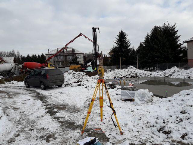 Żółty teodolit na trójnogu ustawiony na zaśnieżonym placu budowy z wylanym fundamentem, w tle betoniarka, pompa do betonu i koparka.