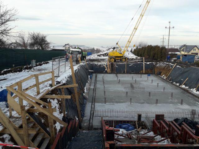 Wykop pod fundamenty domu jednorodzinnego z widocznym zbrojeniem, częściowo zalany betonem, z żółtym dźwigiem na krawędzi wykopu i drewnianymi schodami prowadzącymi w dół, zimowa sceneria w tle.