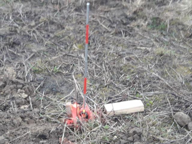 Czerwono-biały tyczka geodezyjna wbita w ziemię z widocznym drewnianym palikiem obok, na tle suchej trawy i ziemi.