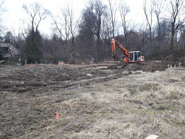 Pomarańczowa koparka Hitachi na placu budowy z wykopanymi fundamentami i drewnianymi stemplami, widok na zalesiony teren.