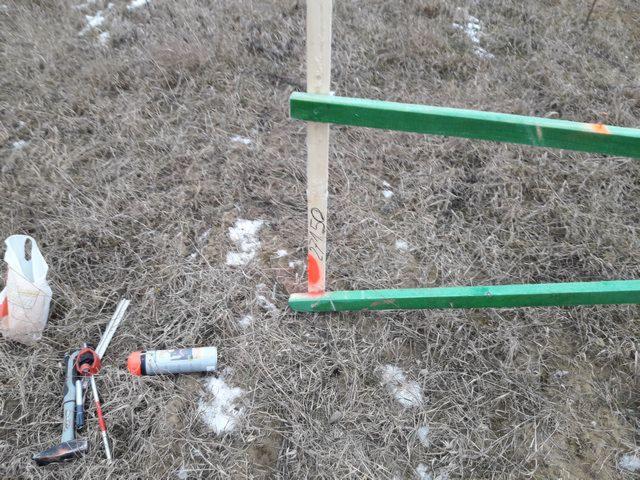 Wytyczenie punktu geodezyjnego: drewniany palik z numerem 2750, zielona poprzeczka, spray z czerwoną nakrętką i klucze leżą na ziemi pokrytej suchą trawą i resztkami śniegu.