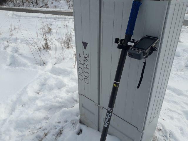 Geodezyjny tyczka z odbiornikiem GPS marki Trimble oparta o szarą skrzynkę z naniesionymi pomiarami wysokościowymi, zasypana śniegiem.