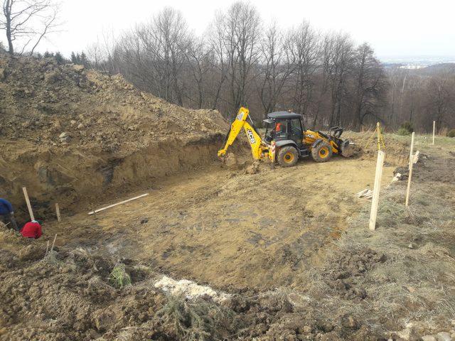 Żółta koparko-ładowarka JCB podczas prac ziemnych na nierównym terenie z wykopem, widoczne drewniane pale wyznaczające granice, w tle drzewa i odległy krajobraz miejski.