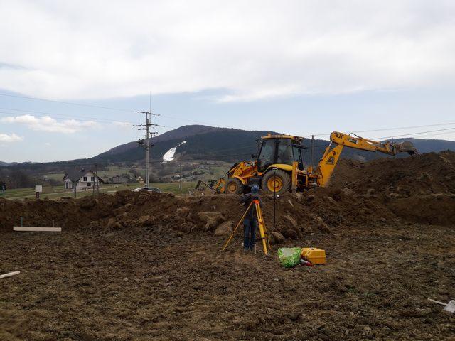 Prace ziemne w Krakowie: Żółta koparka JCB na placu budowy, geodeta z niwelatorem optycznym, w tle góra i zabudowania.