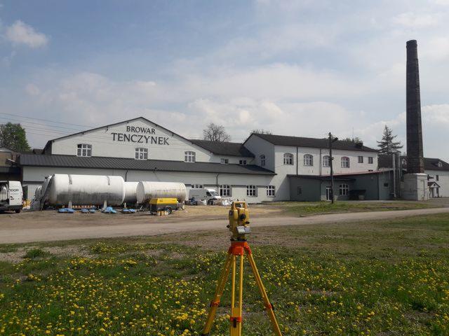 Żółty teodolit na trójnogu ustawiony na łące z mniszkami lekarskimi, w tle budynek Browaru Tenczynek z wysokim kominem. Widoczne zbiorniki i pojazdy.