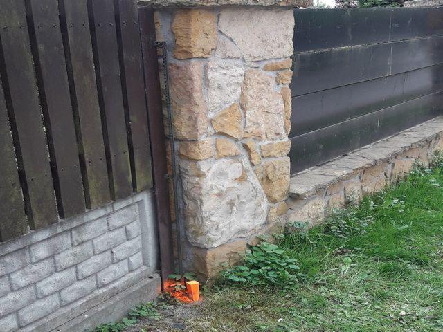 Fragment ogrodzenia z drewnianymi sztachetami po lewej, kamiennym słupkiem pośrodku i metalową bramą po prawej stronie, z pomarańczowym palikiem geodezyjnym u podstawy słupka.