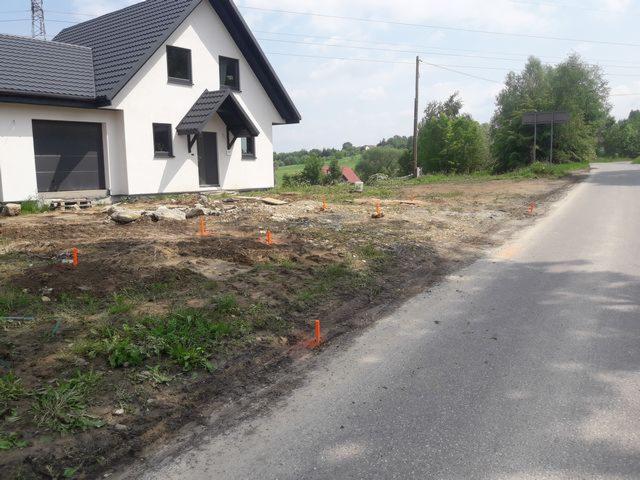 Wytyczanie fundamentów domu jednorodzinnego przy drodze, widoczne pomarańczowe paliki geodezyjne w gruncie.