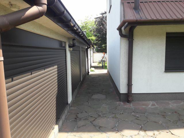 Widok z poziomu gruntu na budynek z brązowymi roletami zewnętrznymi i brązowymi rynnami, widoczna kamienna ścieżka i fragment osoby w oddali.