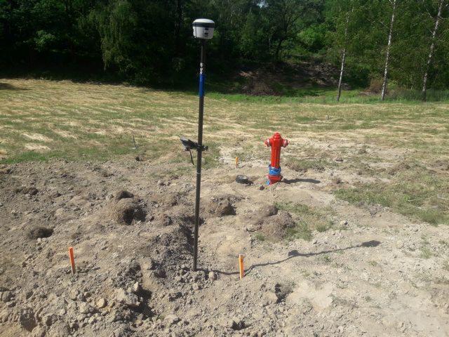 Pomiar terenu z użyciem odbiornika GNSS RTK na tyczce w pobliżu hydrantu, widoczne pomarańczowe paliki wyznaczające punkty pomiarowe.