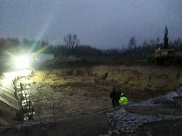 Prace geodezyjne na placu budowy po zmroku, widoczny oświetlony wykop, dwaj geodeci w kurtkach oraz koparka CAT w tle.