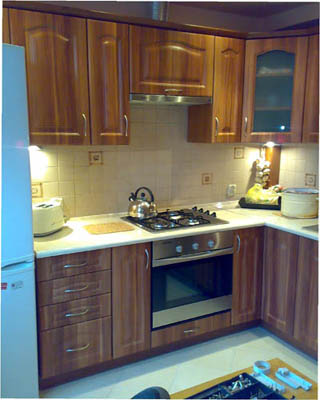 Drewniana zabudowa kuchenna z beżowym blatem, widoczna kuchenka gazowa z czajnikiem, piekarnik, szafki wiszące i stojące, płytki na ścianie, lodówka po lewej stronie.