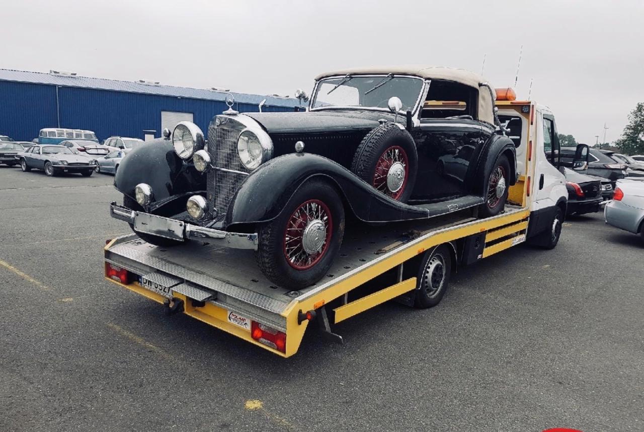 Czarny zabytkowy kabriolet Mercedes na żółtej lawecie pomocy drogowej, z czerwonymi felgami i beżowym dachem, na parkingu z innymi samochodami w tle.
