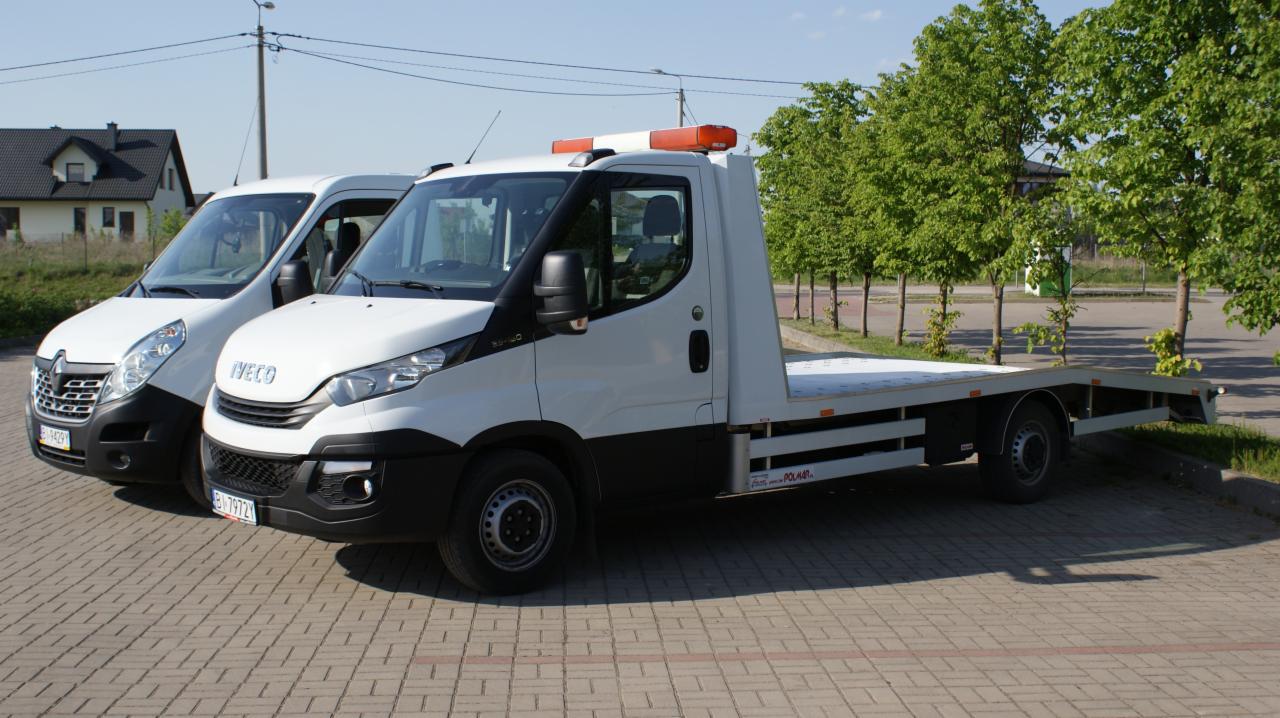 Dwa białe pojazdy: Renault Master i Iveco Daily laweta na kostce brukowej, z domem i drzewami w tle.
