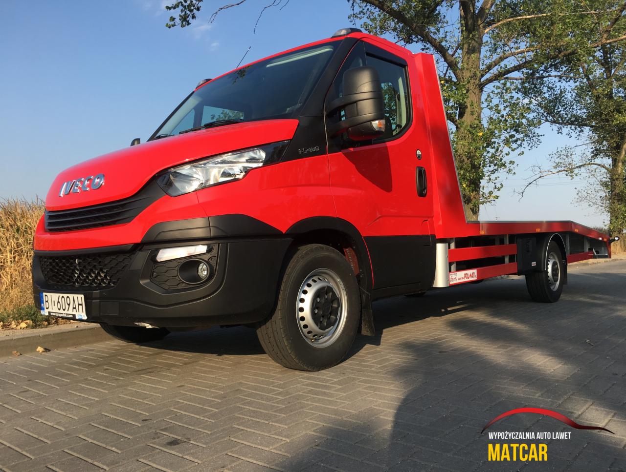 Czerwona laweta Iveco z tablicą rejestracyjną BI 609AH zaparkowana na kostce brukowej, widok z niskiej perspektywy, w tle drzewa i pole.