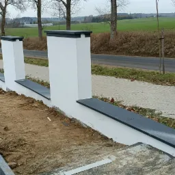 Wznoszenie ogrodzenia z białych słupków z ciemnymi daszkami i podmurówką na posesji w trakcie prac ziemnych.