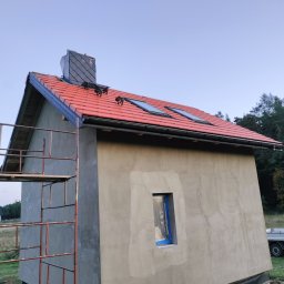 Usługi Blacharsko-Dekarskie Łukasz Siwkowski - Niedawno ukończony budynek z czerwonym dachem, dwoma oknami dachowymi, kominem z szarej dachówki łupkowej i nowymi rynnami, częściowo zakryty rusztowaniem, na tle zalesionego wzgórza.