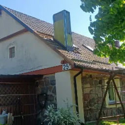 Dom jednorodzinny z brązowym dachem, na którym widoczny jest komin w trakcie montażu, zabezpieczony żółtą izolacją. Część dachu jest odsłonięta, a w tle błękitne niebo i fragment drzewa.