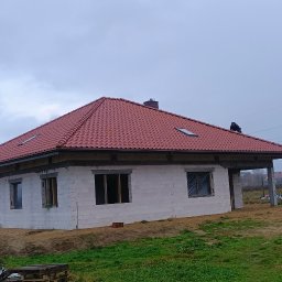 Usługi Blacharsko-Dekarskie Łukasz Siwkowski - Dom w trakcie budowy z nowym, czerwonym dachem z dachówki ceramicznej; widoczny dekarz na dachu oraz częściowo wykończona elewacja z białego bloczka.